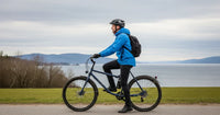 Un homme en vélo, portant un casque et un sac à dos, pédale le long d'une route près d'un lac ou d'une mer, sous un ciel nuageux, illustrant une activité de plein air saine.