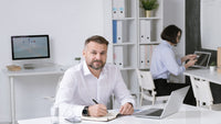 Un homme d'affaires concentré, assis à un bureau moderne avec un ordinateur portable, des documents et un verre d'eau, dans un environnement de bureau épuré, illustrant la gestion du stress entrepreneurial.